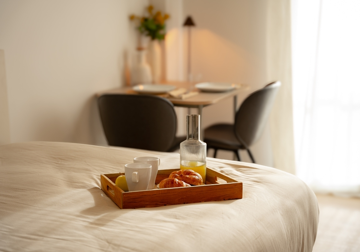 Bandeja con desayuno servido sobre una cama en un estudio de Calido Living, con mesa de comedor al fondo.